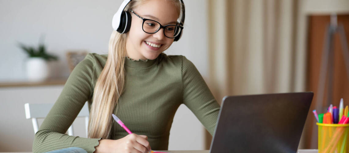 Positive school girl in cute glasses and wireless headset studying from home in front of laptop, looking at computer screen and taking notes, copy space. E-education, online leraning concept